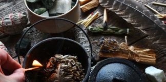 A close up image of a smoldering white sage smudge bundle and healing crystal on a dark wooden table.