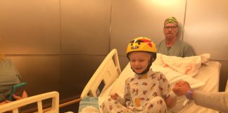 A young boy smiling in a hospitla bed, wearing a protective helmet after brain surgery.